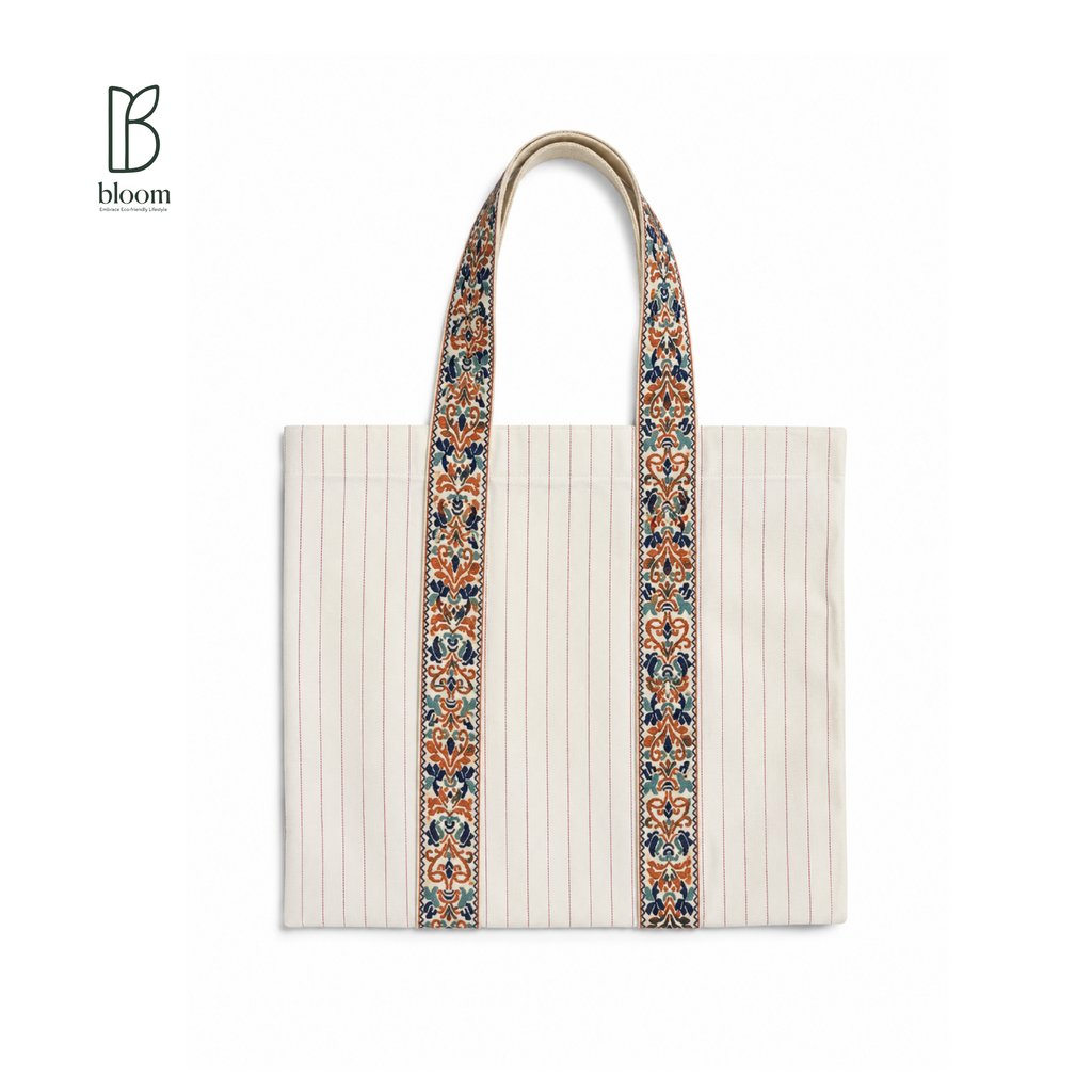 A cream-colored canvas tote bag with vertical pinstripes and ornate, multicolored patterned shoulder straps, shown on a plain white background with a 'bloom' brand logo in the top-left corner.